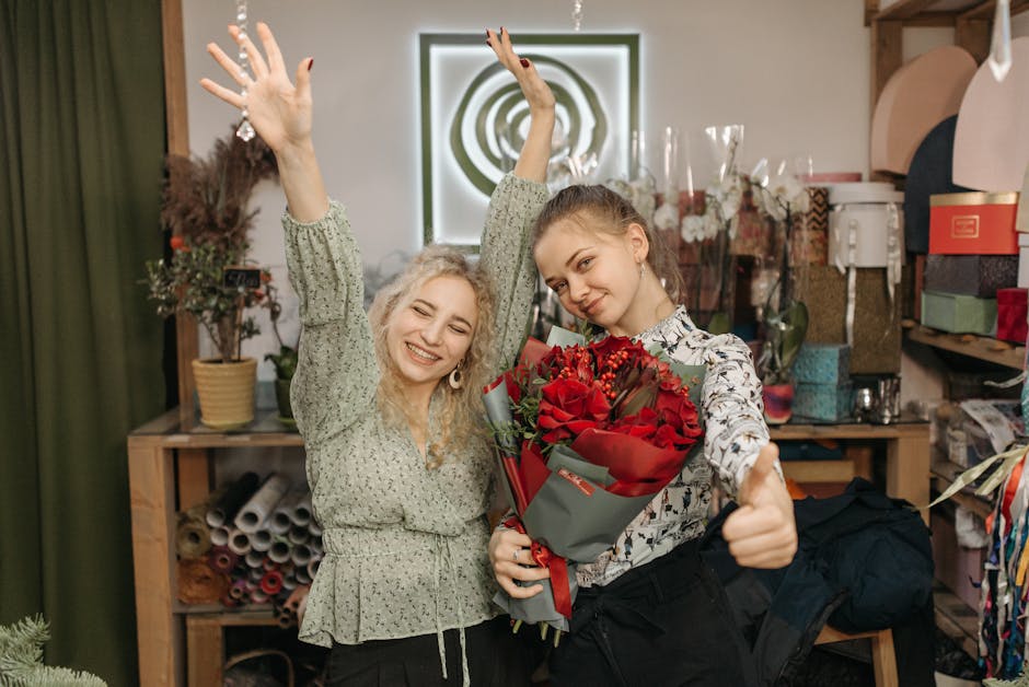 Two women inside a flower shop with wooden shelving and potted plants in the background. The woman on the left has curly blonde hair and is wearing a light green blouse with a floral pattern, raising