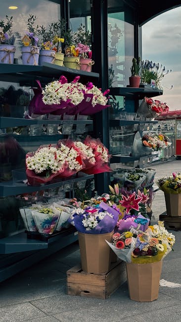 A display of floral bouquets and potted plants arranged on metal shelves outside a flower shop, showcasing a variety of fresh flowers including pink lilies, white chrysanthemums, purple irises, and mi