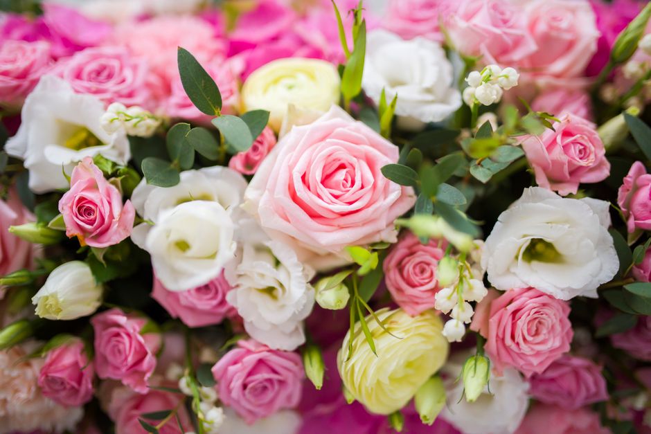 A close-up view of a floral arrangement featuring a variety of fresh roses, lisianthus, and small white blossoms, predominantly in soft pink, white, and pale yellow hues. The bouquet includes lush gre
