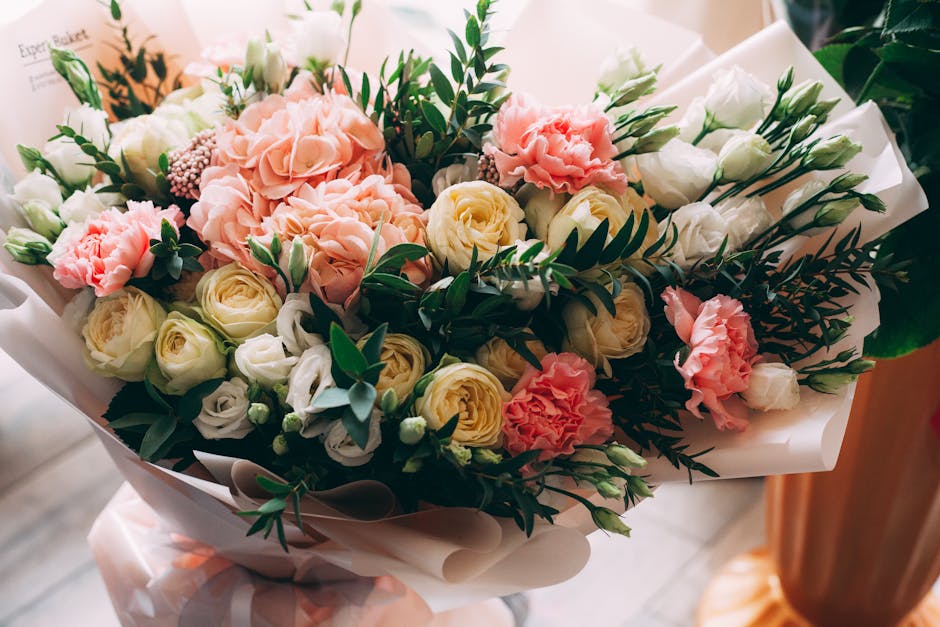 A bridal bouquet arrangement featuring a mix of soft pastel-colored flowers including pale yellow roses, white lisianthus, and pink carnations, accented with lush green foliage and eucalyptus leaves.