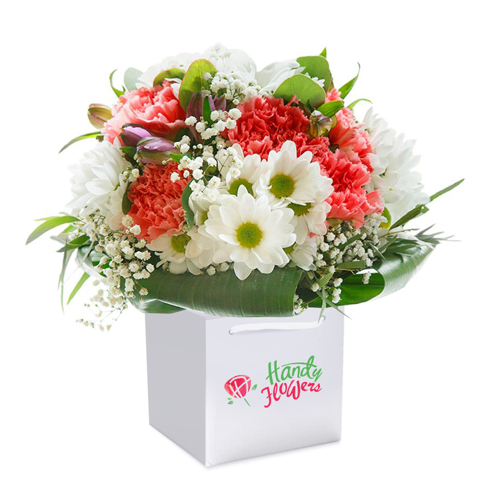 Beautifully presented within a classic white square gift bag, this floral arrangement exudes the natural charm of Downe, mirroring the wild beauty found along its bridleways and gardens. Rich coral and vivid red carnations, known for their luxuriant, frilly petals, form the heart of the bouquet, surrounded by crisp, white daisies crowned with bright lime-green centers. Wispy clusters of gypsophila (baby's breath) intersperse throughout the arrangement, softening the profile with their light, airy presence. The composition is cradled in generous sweeps of deep, glossy green foliage, its texture and shine reminiscent of Downe's hedgerows after a summer rain. On the left, discrete hints of purple-tinted buds emerge, suggesting mystery and the quiet anticipation of new floral life. The bouquet's rounded shape is both structured and abundant-crafted to communicate freshness and the spirit of sharing. Sturdy white ribbon handles lend the gift bag practical elegance, suited for easy delivery to a friend's home on Main Road or a celebration in one of Downe's scenic gardens. Against a pure white studio backdrop, every petal, stem, and leaf is revealed in sharp, vibrant detail, summoning memories of the countryside's tranquil calm. This bouquet is ideal for birthdays, anniversaries, or simply letting someone know you care-each bloom meticulously placed by Downe Flowers to reflect the village's rich floral heritage and neighbourly warmth.